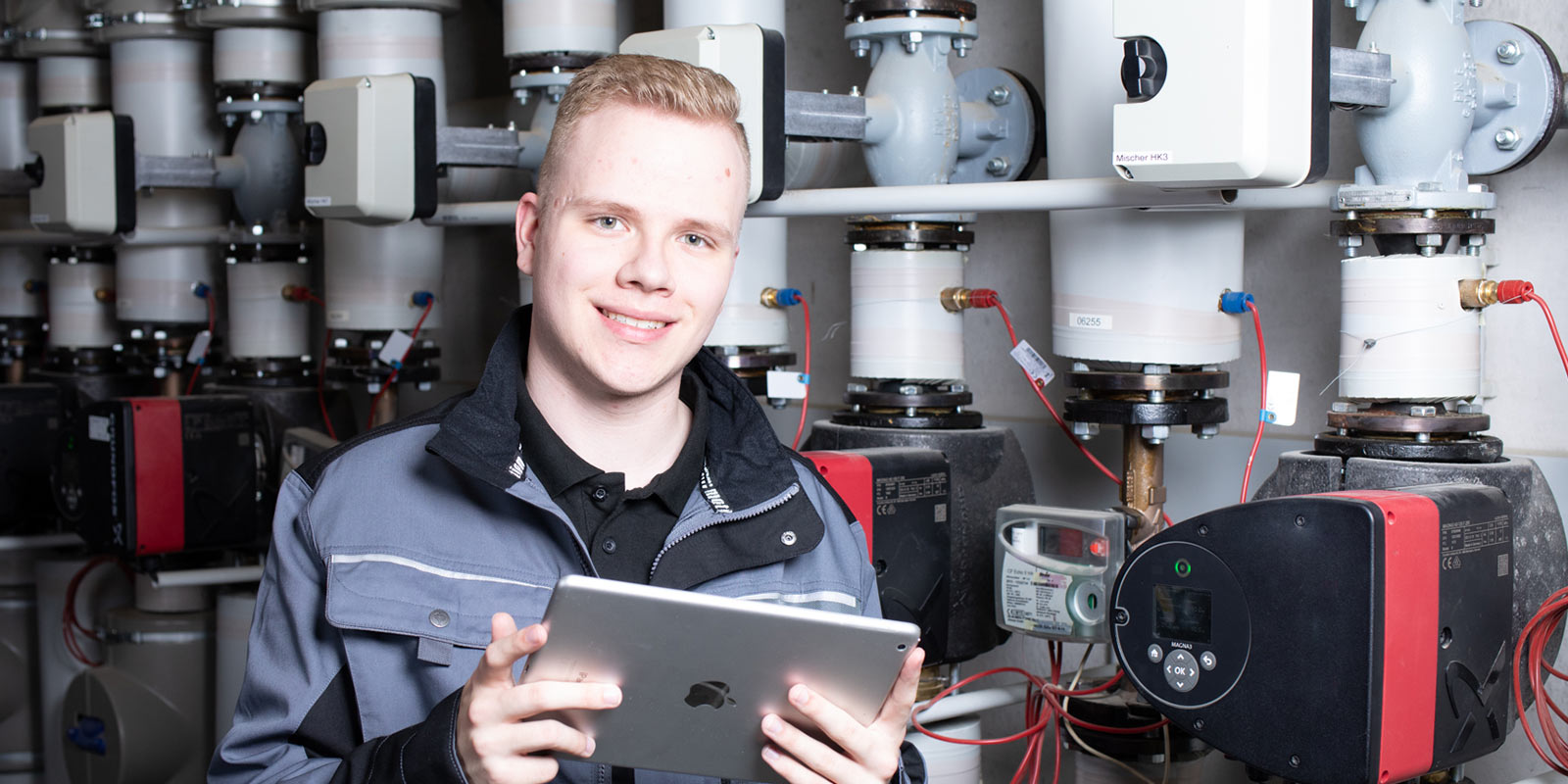 Ein junger Anlagenmechaniker vor einer Heizungsanlage, der ein Tablet hält 