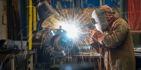 Ein Schweißer arbeitet konzentriert an einem Metallstück, während Funken in alle Richtungen sprühen und eine helle Lichtquelle entsteht.