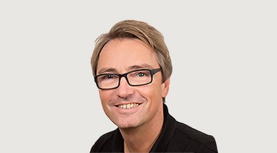 Ein lächelnder Mann mit Brille und blonden Haaren posiert vor einem neutralen Hintergrund, strahlt Freundlichkeit aus.