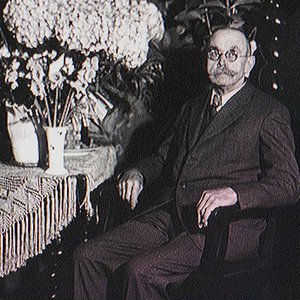 Ein älterer Mann mit Brille sitzt entspannt in einem Stuhl, umgeben von üppigen Blumenarrangements auf einem Tisch.